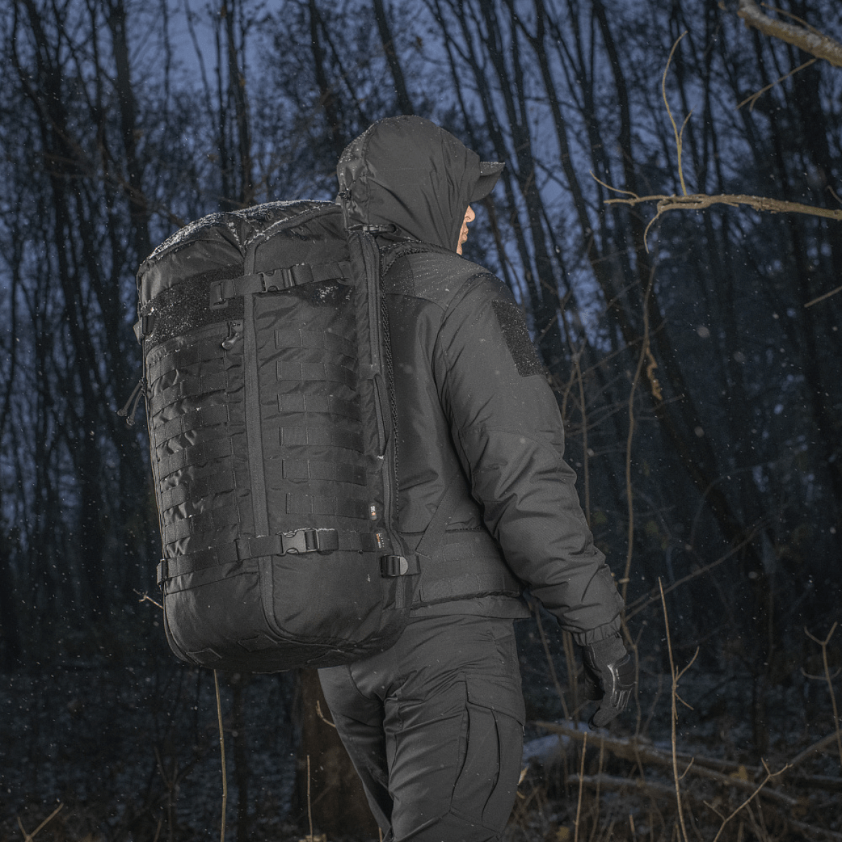 M-Tac Rygsæk Gen.II Elite Large, henkilö talvitakissa metsässä, esittelee kestävyyttä ja toiminnallisuutta kylmässä säässä.