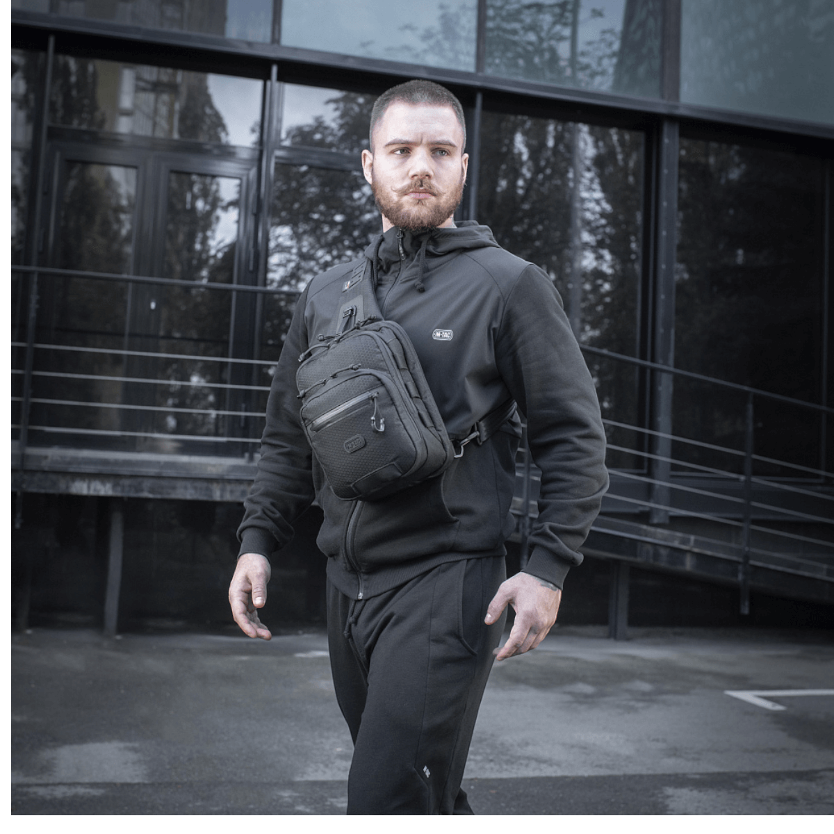 Mies M-Tac Cross Taske Slim Elite Hex -laukku olallaan, pukeutunut mustiin vaatteisiin modernin rakennuksen edessä.
