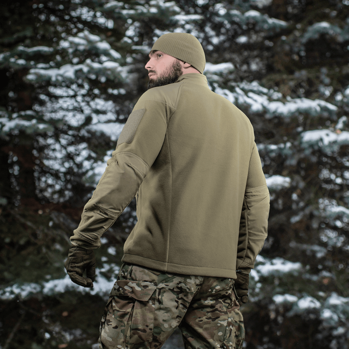 Mies M-Tac Combat Fleece Polartec -takissa metsässä, puissa lunta, takaa nähtynä, näyttää toimivan designin ja materiaalit.