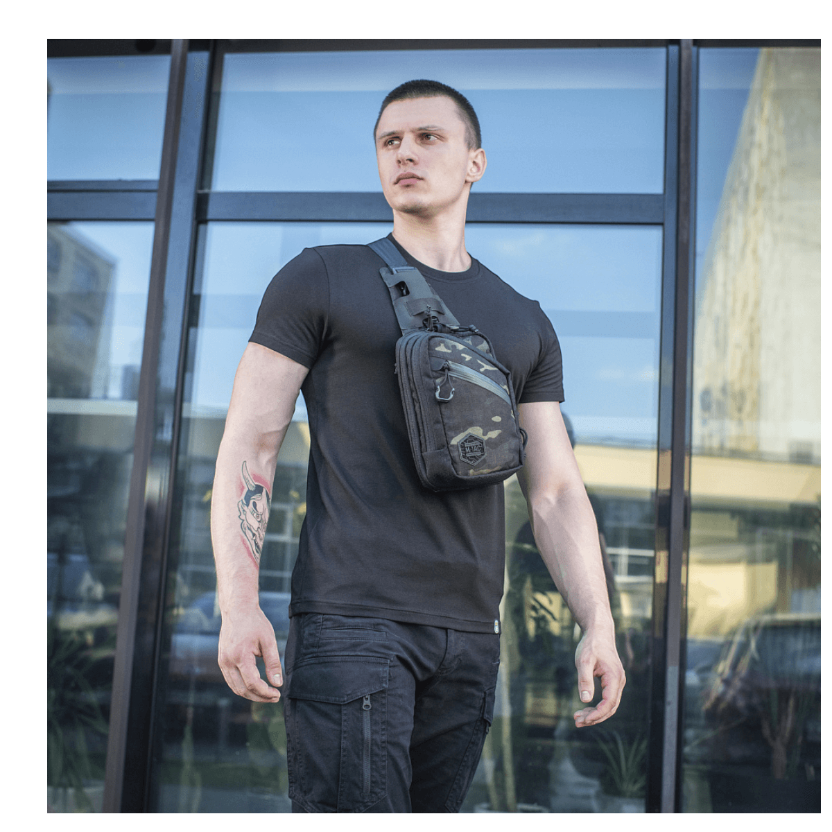 Mies M-Tac Sling Pistoltaske Elite Hex -laukulla lasirakennuksen edessä, näyttää monipuolisuutta ja tyyliä.