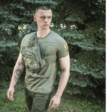 Mies vihreällä M-Tac Sling Pistoltaske Elite Hex -laukkulla, valmistettu vahvasta Cordurasta, säädettävä hihna metsäalueella.