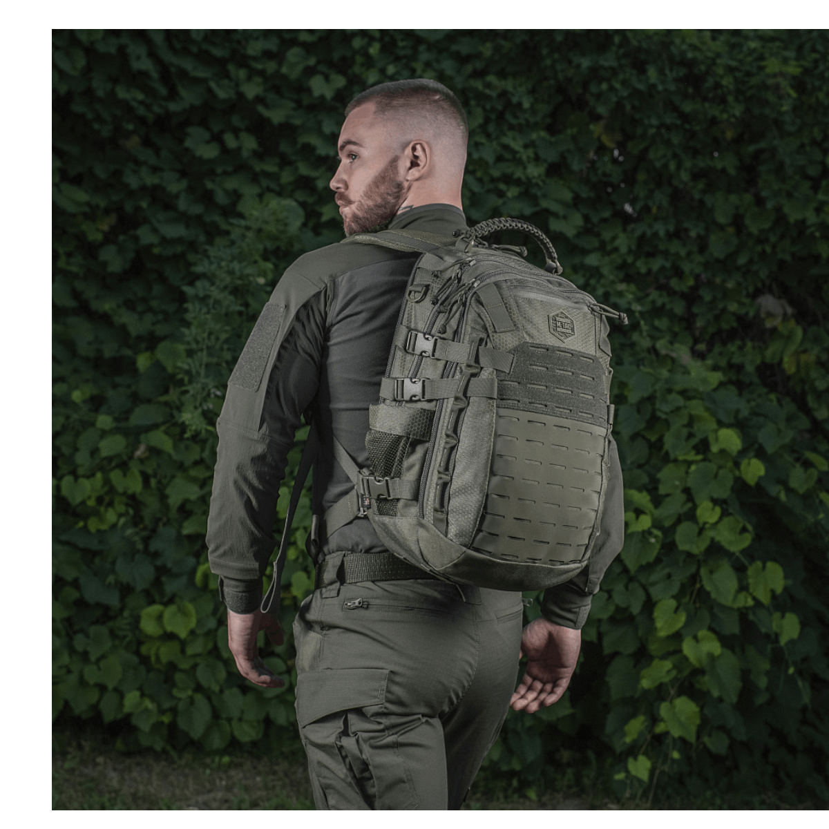 Henkilö M-Tac Mission Rygsæk Elite Hex -repun kanssa, taustalla vihreää kasvillisuutta.