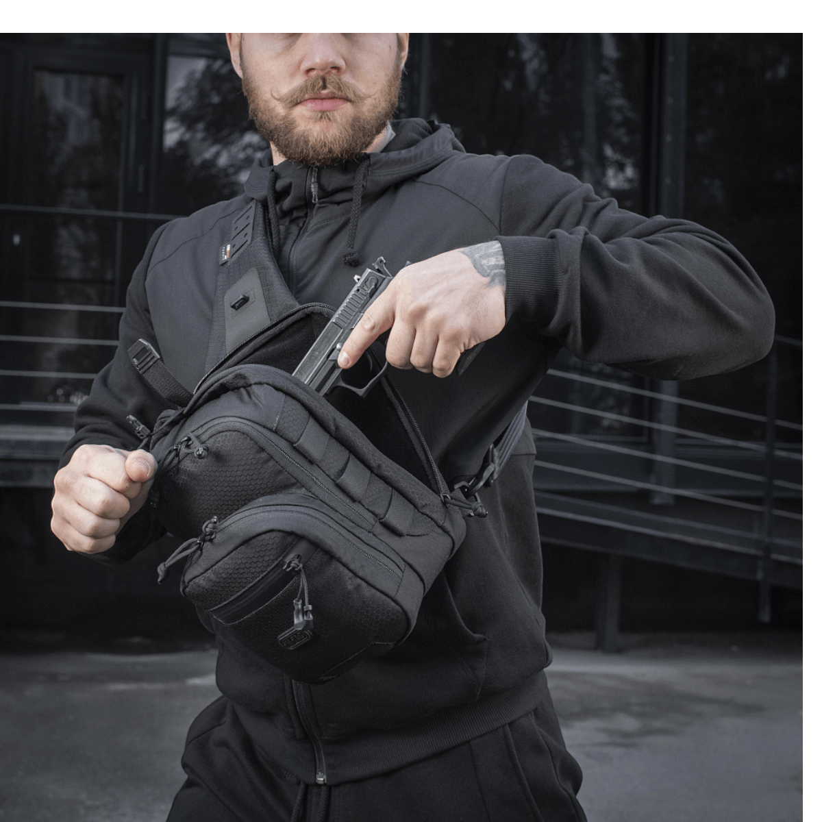 Mies käyttää M-Tac Cross Taske Slim Elite Hex -laukkua mustana Cordura-materiaaleilla, vetoketjuilla ja YKK- ja Woojin-soljilla, ulkoilmassa.