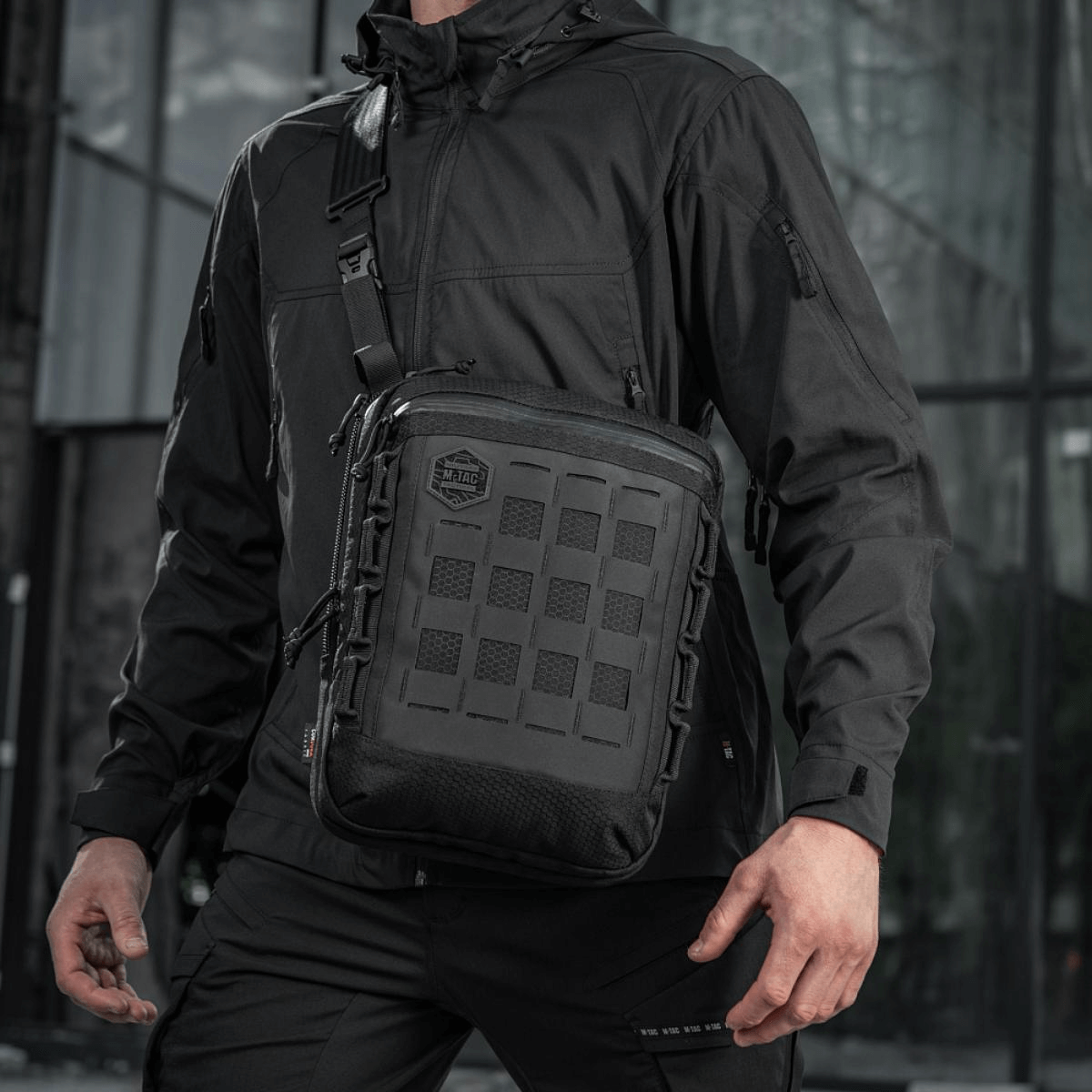 Mies mustassa takissa kantaa M-Tac Tablet Bag Laser Cut Hex -laukkua, tukeva ja käytännöllinen EDC- ja piilotuskantolaukku.