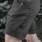 Mies pukeutuneena M-Tac Shorts Aggressor Shortiin, mustat polvipituiset shortsit monilla taskuilla, ihanteelliset taktiseen käyttöön lämpimässä säässä. Valmistettu polycotton Flex rip-stop -materiaalista.