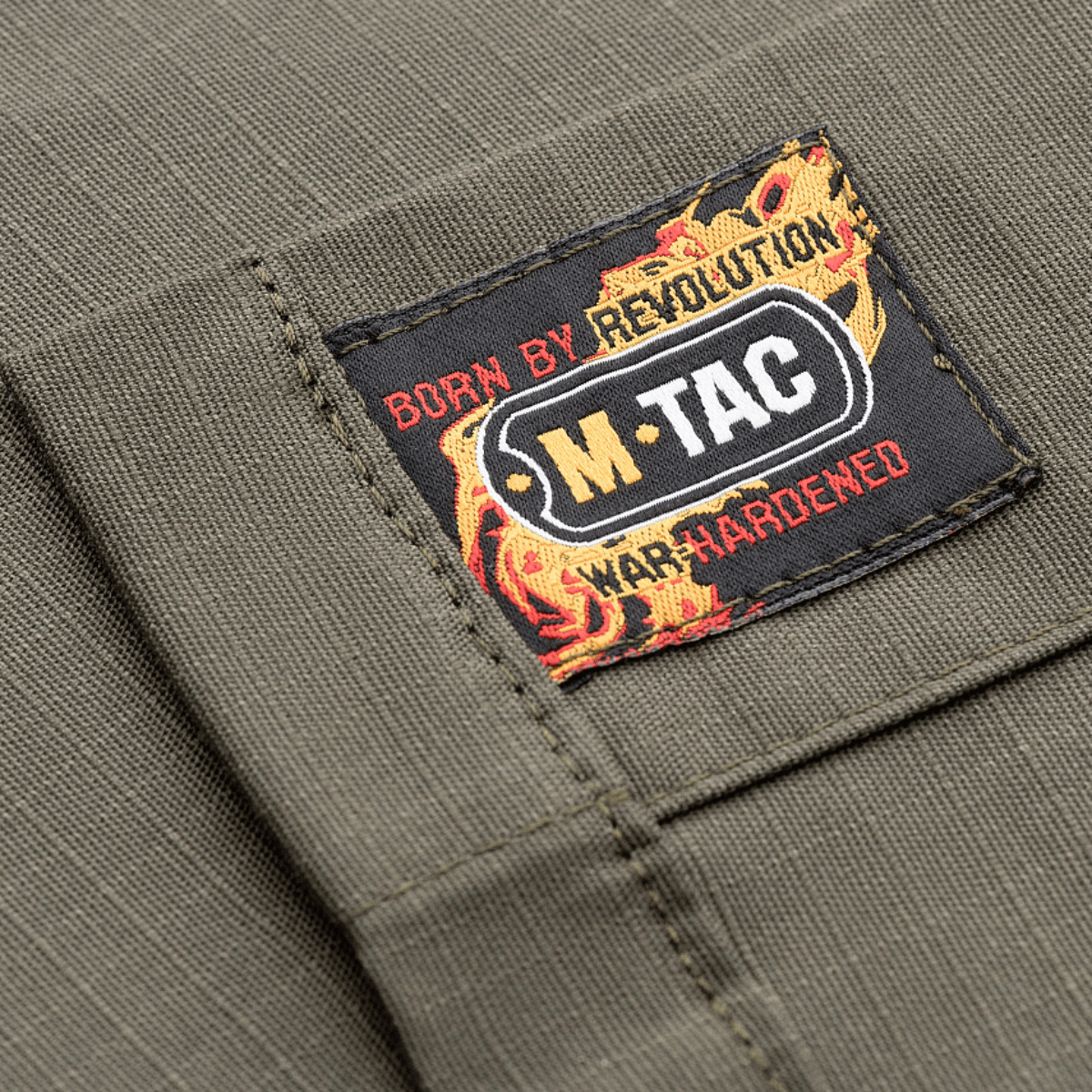 M-Tac Patrol Flex -takin etiketti tekstillä "Born by Revolution" ja "War Hardened", harmaa väri.