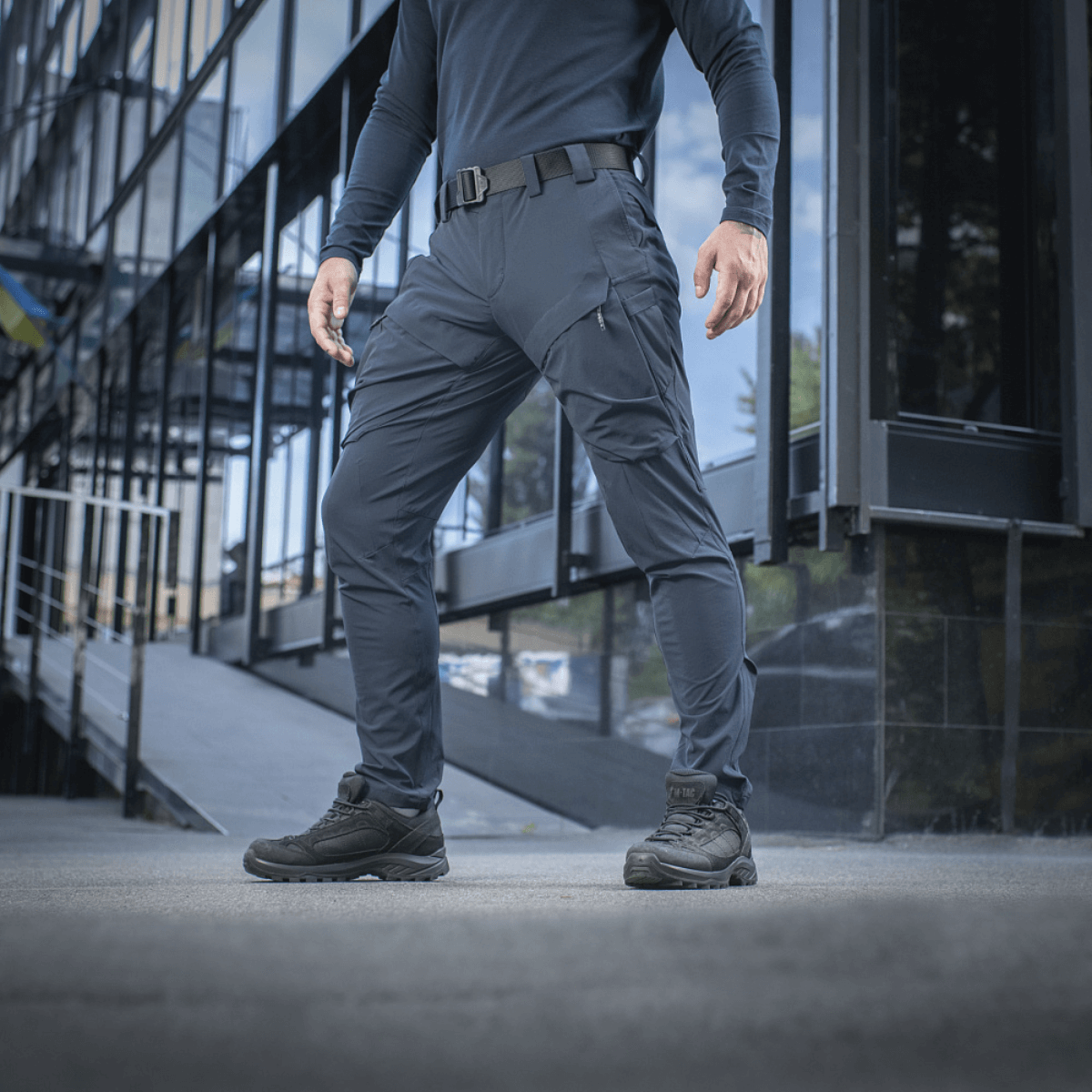 Mies pukeutuneena M-Tac Rubicon Flex -housuihin innovatiivisella cargo-muotoilulla, sopivat harjoitteluun, työhön ja vapaa-aikaan, näytetty modernin rakennuksen taustalla.