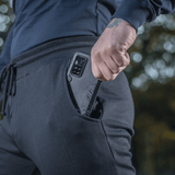 M-Tac Stealth Cotton -housut matkapuhelin sivutaskussa, korostaa mukavuutta ja toiminnallisuutta, sopii urheiluun ja arkeen.