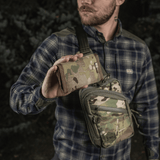 Mies pitelee M-Tac Pung Elite Gen.II:tä maastokuvioisena, valmistettu Cordura 500D:stä ja varustettu YKK-vetoketjuilla, metsäympäristössä.