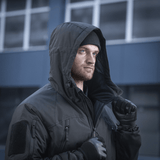 Mies käyttää M-Tac Alpha Gen.III Pro talvitakkia hupun ollessa päällä, mukava lämpötiloihin jopa -20°C, modernin rakennuksen taustalla.