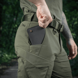 Mies pukeutuneena M-Tac Aggressor Summer Flex Shorts -shortseihin metsässä, käsi taskussa näyttää puhelimen säilytyksen. Tuoteavainsana: M-Tac Aggressor Summer Flex Shorts.