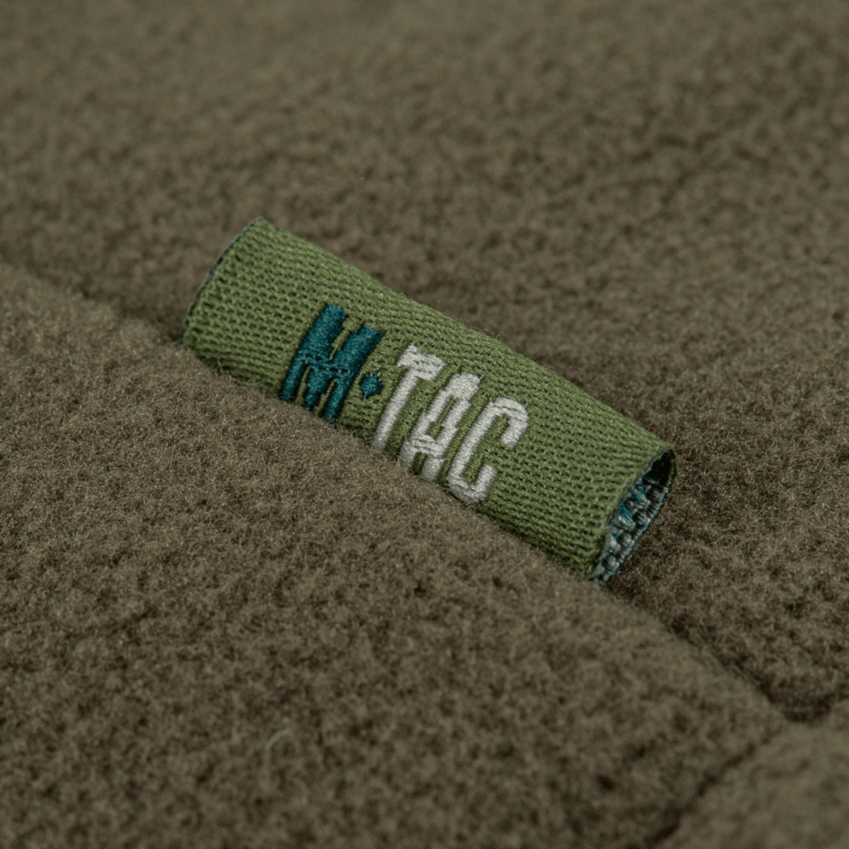 M-Tac Combat Fleece Polartec -takki, fokusoitu merkki hihassa, näyttää tekstuurin ja brändilogon yksityiskohdat.