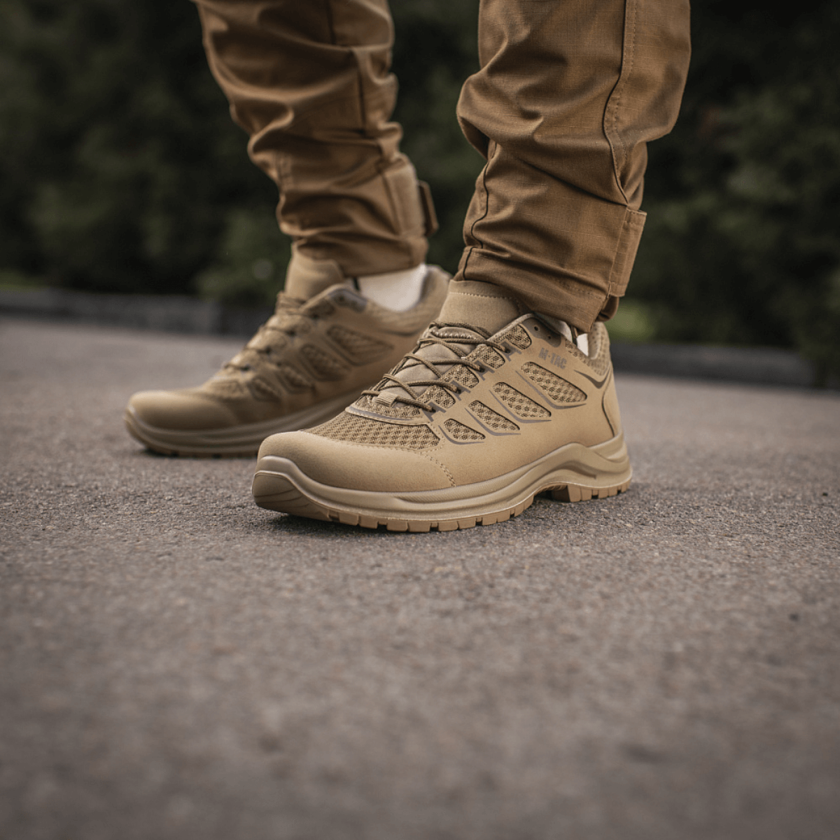 M-Tac Tactical Sneakers IVA jalassa, sopii kesäaktiviteetteihin ja vaellukseen, esitetty asfaltilla vihreää taustaa vasten.