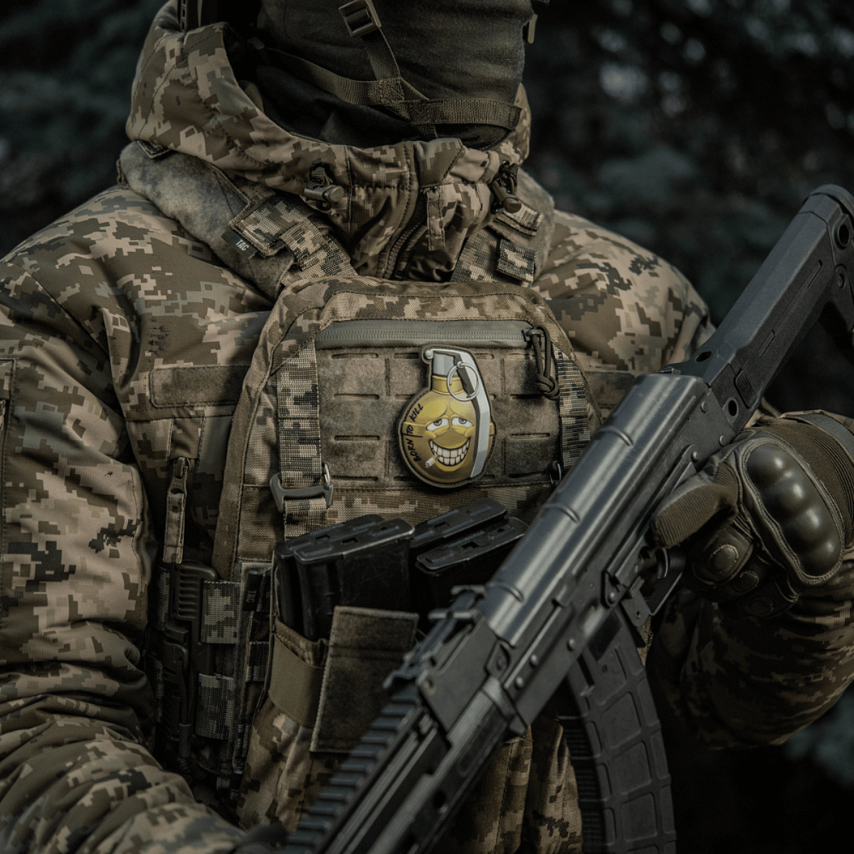 Sotilas maastokuvioisessa takissa kantaa M-Tac Patch M67 Kranaatti 3D PVC:tä liivissä, humoristinen emoji-kuvio, taktinen varustus.