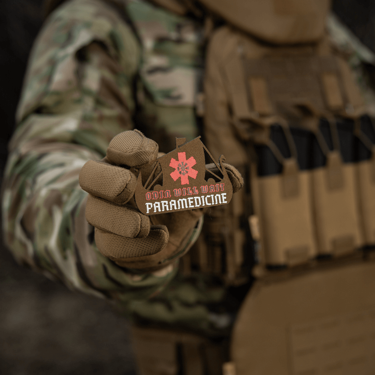 M-Tac Patch Viking Evac sotilaan kädessä, näyttää 'Odin will wait paramedicine' -aiheen. Maastotakki ja taktinen liivi taustalla.