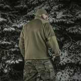 Mies pukeutuneena M-Tac Battle Fleece Polartec -takkiin lumisessa metsässä, nähtynä takaa anatomisella istuvuudella ja maastohousuilla.