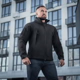 Mies pukeutuneena M-Tac Soft Shell -takkiin seisoo modernin rakennuksen edessä, näyttää takin sääsuojaus- ja eristysominaisuuksia.