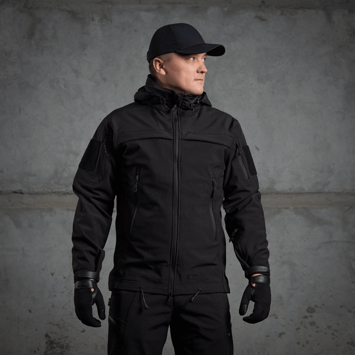 Mies pukeutuneena M-Tac Soft Shell Police Jakkeen, musta takki säädettävin yksityiskohdin ja sisäinen fleece, suunniteltu tuulta ja säätä vastaan.