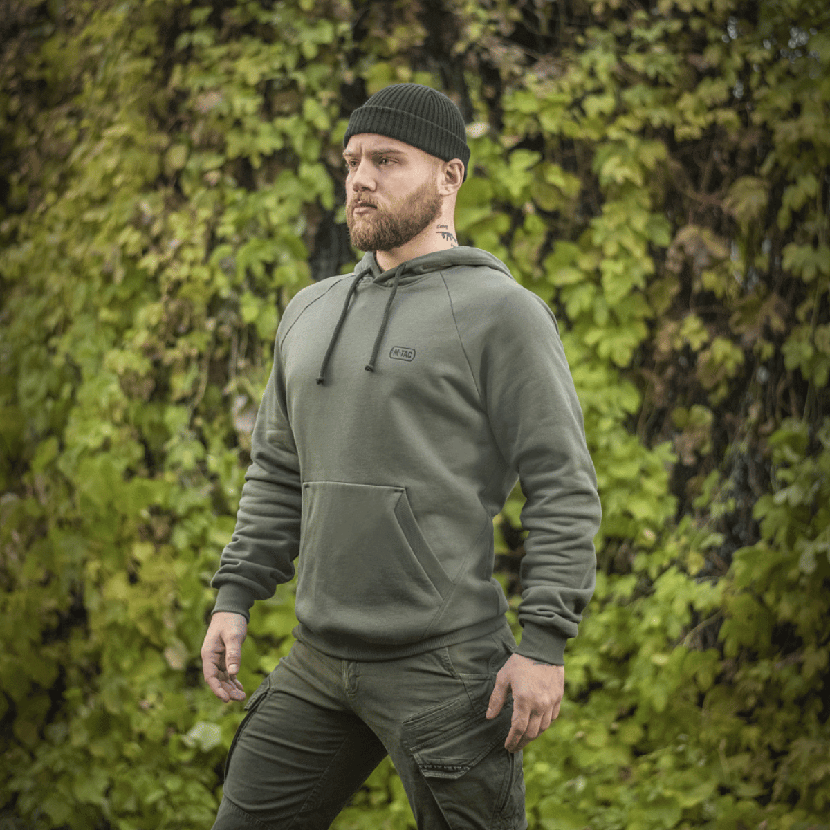 Mies vihreässä M-Tac Cotton Raglan -hupparissa ulkona lehtipeitteisen taustan edessä, rento ja mukava tyyli.