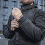 Mies pukeutuneena M-Tac Rubicon -takkiin Gen. II, kestävä ja lämmin bomber-takki, säätää hihoja modernin rakennuksen edessä